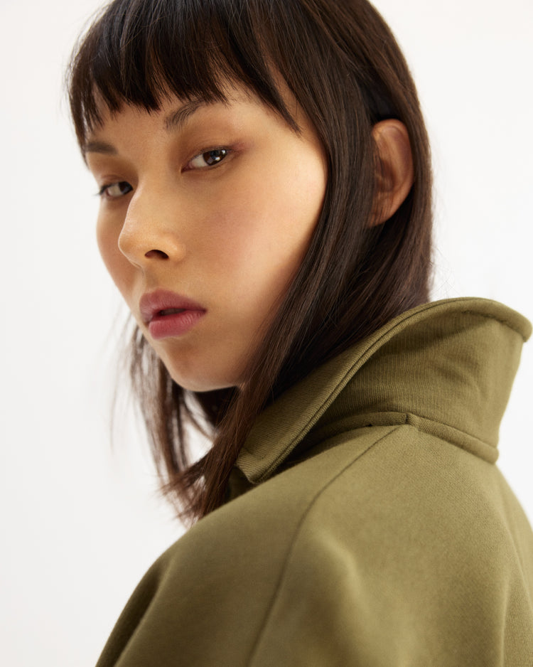 A woman with straight dark hair and bangs looks over her shoulder, wearing the Arlo Mott Elliott Polo Pullover in Olive with the collar turned up against a plain light background.