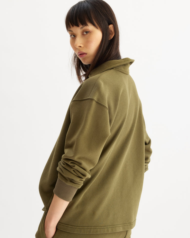 A woman with straight dark hair and bangs, turned slightly away and looking over her shoulder, models the Elliott Polo Pullover in Olive by Arlo Mott with sleeves pushed up against a plain light background.