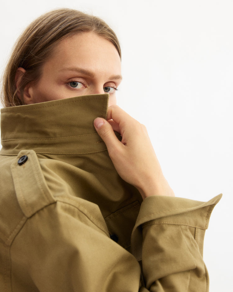 A person with light brown hair partially covers their face with the high collar of the Arlo Mott Henry Short Shirt Jacket in Khaki, looking directly at the camera against a plain white background.
