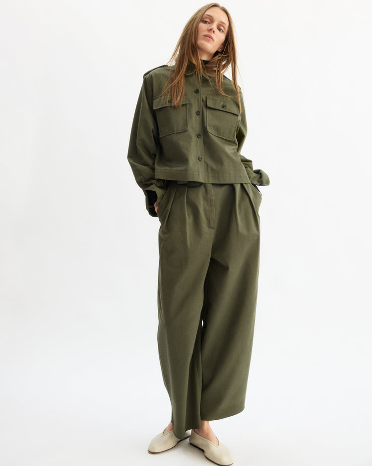A person with long hair wears the Arlo Mott Henry Short Shirt Jacket in Olive, featuring utility details and large pockets, paired with matching wide-leg pants and light slip-on shoes, standing against a plain white background.
