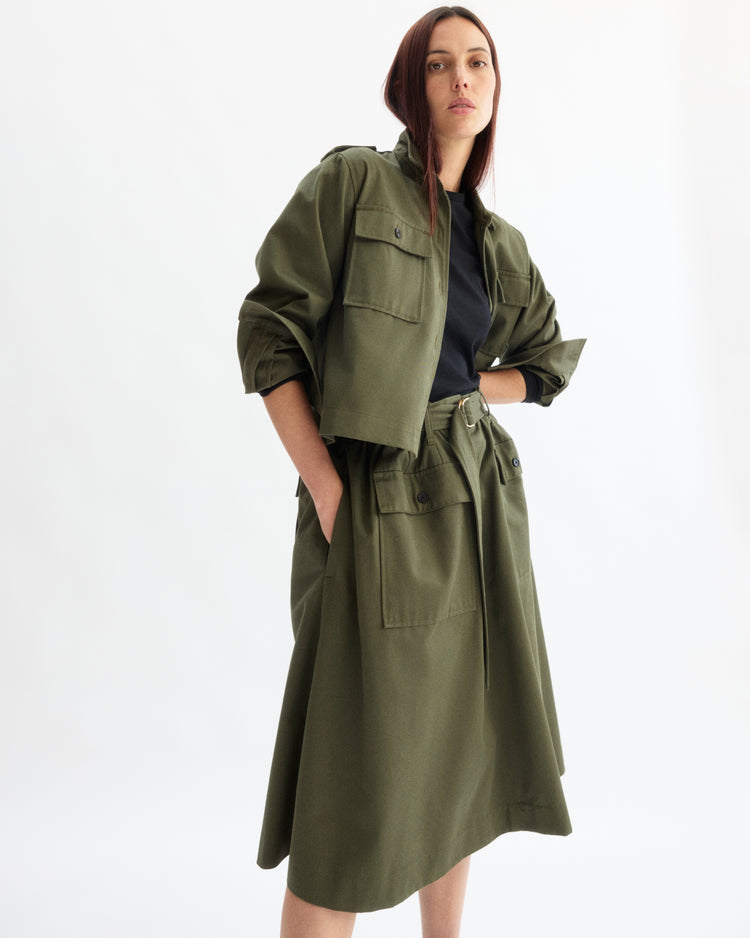 A woman stands with hands in pockets, wearing the Arlo Mott Felix Utility Midi Skirt in Olive and a matching utility jacket over a black top, set against a plain white background.