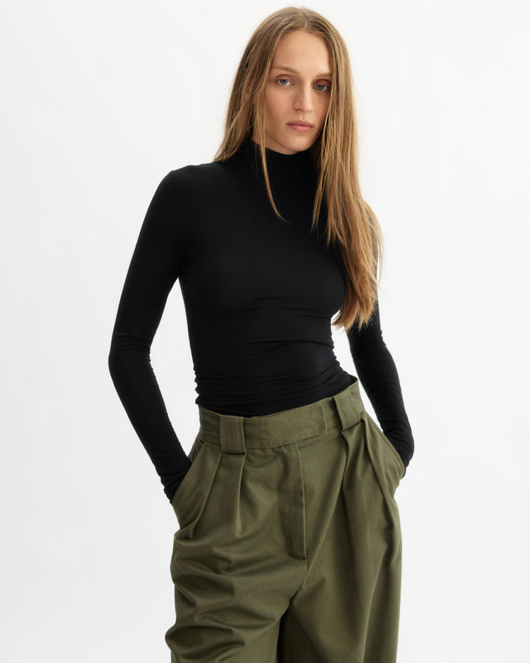 A woman with long, straight hair wears the Arlo Mott Jones Long Sleeve Turtleneck in Black and high-waisted olive green trousers, standing against a white background with her hands in her pockets.