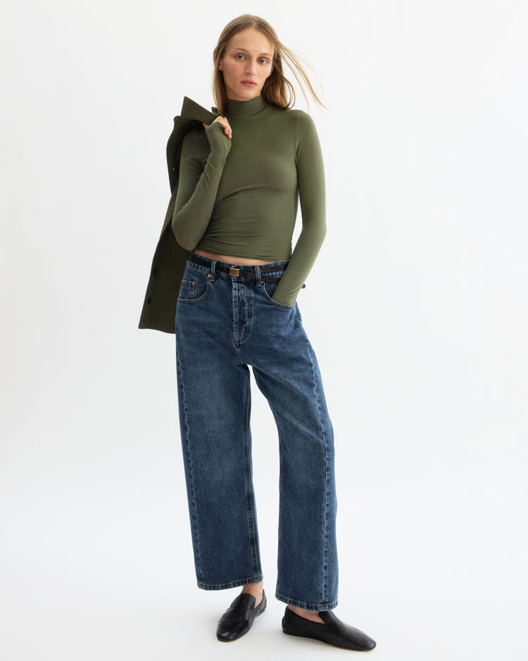 A woman stands against a white background wearing the Arlo Mott Jones Long Sleeve Turtleneck in Olive, loose blue jeans, black slip-on shoes, and a matching green jacket draped over her shoulder.