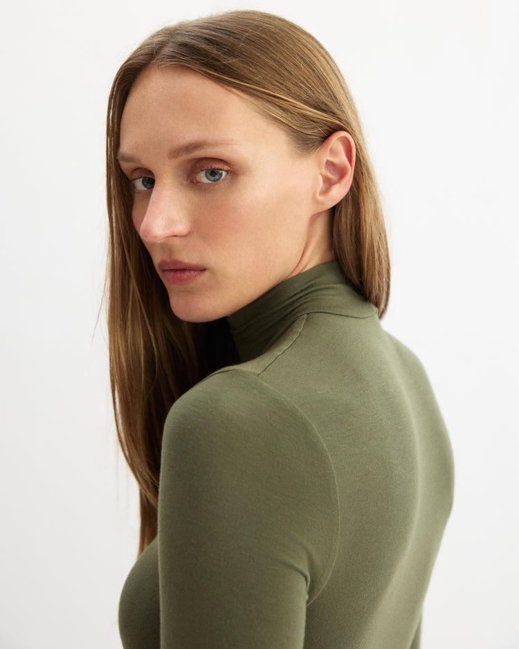 A woman with straight, light brown hair wears the Arlo Mott Jones Long Sleeve Turtleneck in Olive and looks over her shoulder toward the camera against a plain white background.