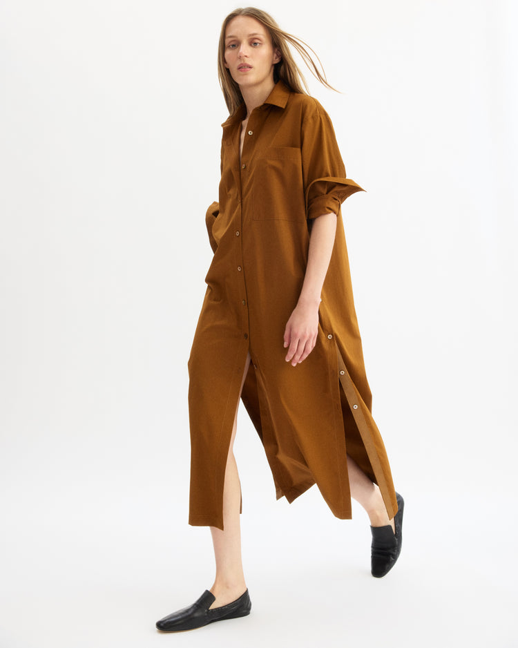A woman walks confidently against a white background wearing the Arlo Mott Lafayette Side Button Maxi Dress in Cinnamon with mother of pearl buttons, side slits, and black slip-on shoes.