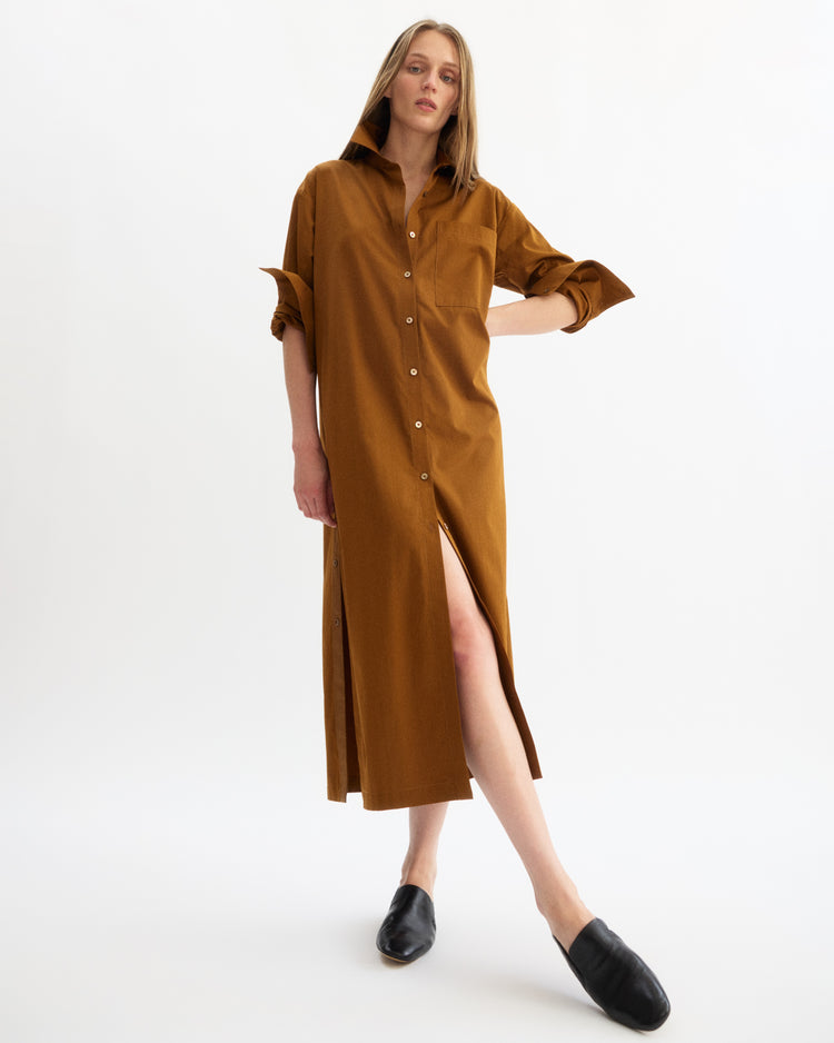 A woman models the Lafayette Side Button Maxi Dress in Cinnamon by Arlo Mott, featuring Portuguese poplin, rolled sleeves, and a high slit. She pairs it with black slip-ons and stands against a plain white background.