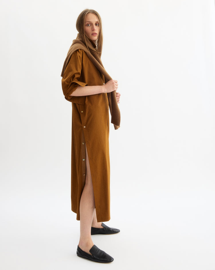 A woman stands sideways against a white background, wearing the Arlo Mott Lafayette Side Button Maxi Dress in Cinnamon with mother of pearl buttons, a matching sweater over her shoulders, and black slip-on shoes.