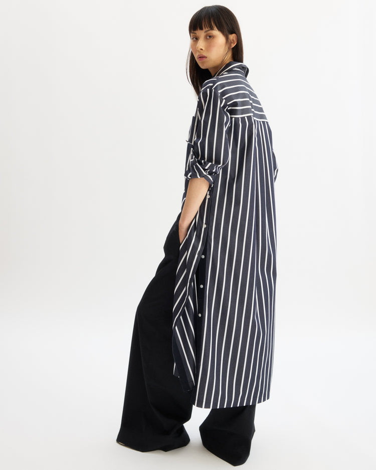 A woman stands in profile against a white background wearing the Arlo Mott Lafayette Side Button Maxi Dress in Navy Stripe, crafted from Portuguese poplin, sleeves rolled up, layered over wide-leg black pants with hands in pockets.