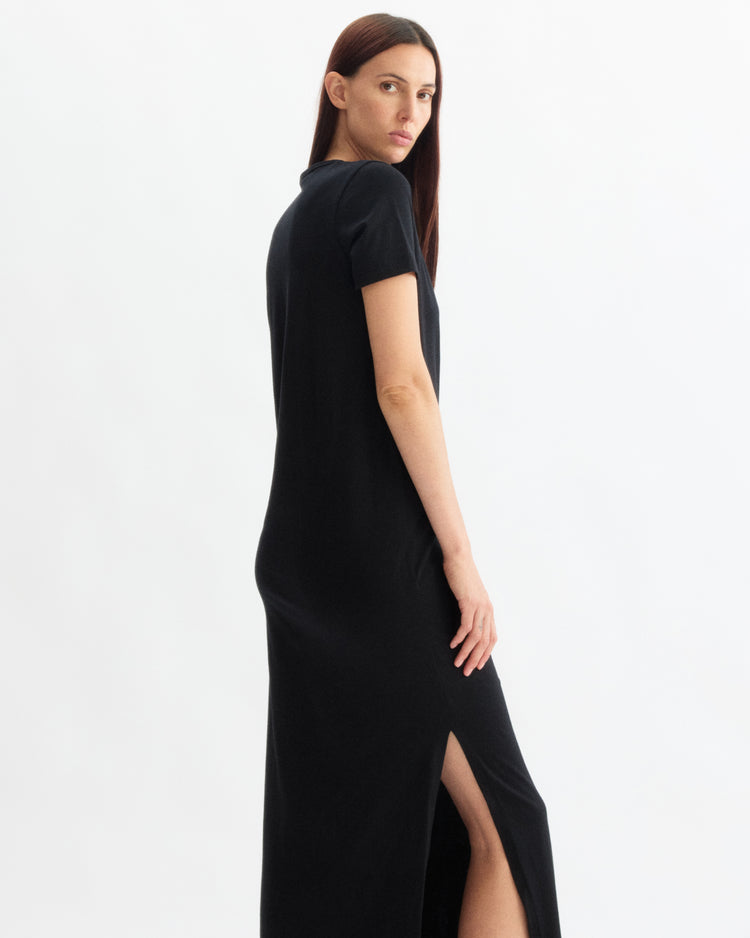 A woman with straight brown hair models the Arlo Mott Hubert Short Sleeve Maxi Dress in Black, featuring a side slit, standing sideways and looking over her shoulder against a plain white background.