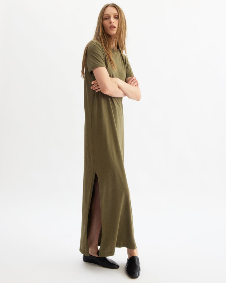 A person stands against a plain white background wearing the Arlo Mott Hubert Short Sleeve Maxi Dress in Olive, paired with black slip-on shoes and sporting long, straight hair.