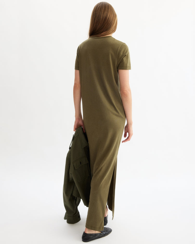 A woman with long hair, facing away, wears the Arlo Mott Hubert Short Sleeve Maxi Dress in Olive and holds a matching jacket in her left hand. She pairs black flats with the dress, standing against a plain white background.