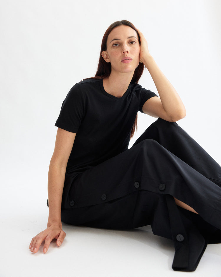 A woman with straight brown hair sits on the floor against a white background, wearing an Arlo Mott black Portuguese cotton t-shirt and Baxter Side Button Pants in Black, resting her head on her hand and looking at the camera.