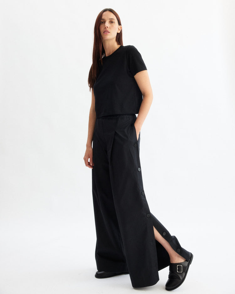 A woman with long brown hair wears a black short-sleeve top and Arlo Mott’s Baxter Side Button Pants in Black, featuring an elastic waist and side buttons, paired with black slip-on shoes. She stands against a plain white background.