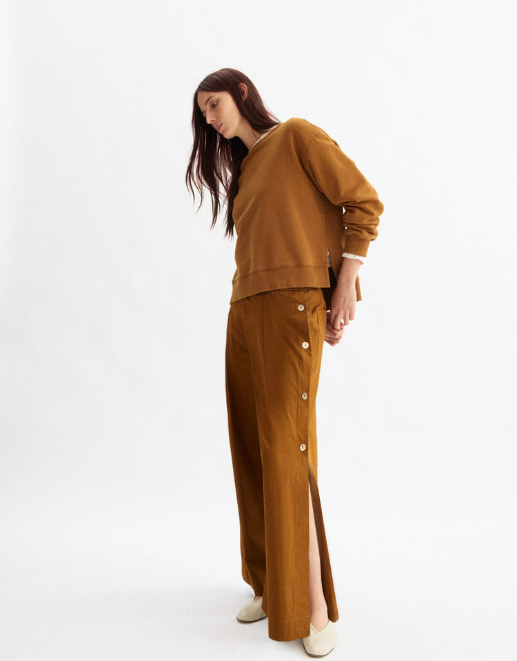 A woman stands against a white background, wearing Arlo Mott's Baxter Side Button Pants in Cinnamon—wide-legged Portuguese cotton pants with side buttons, slits, and an elastic waist—paired with a loose brown sweater and white shoes as she looks down.