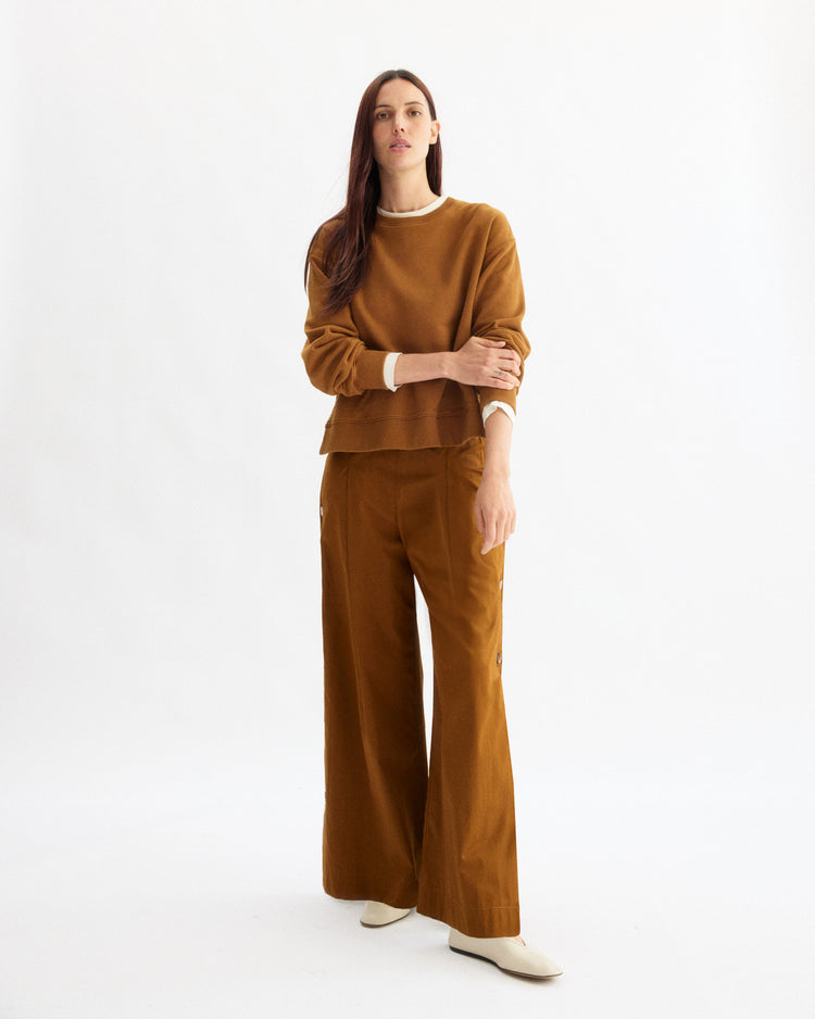 A woman with long brown hair and a neutral expression stands against a white background wearing an Arlo Mott loose brown sweater over a white top, Baxter Side Button Pants in Cinnamon by Arlo Mott, and white shoes.
