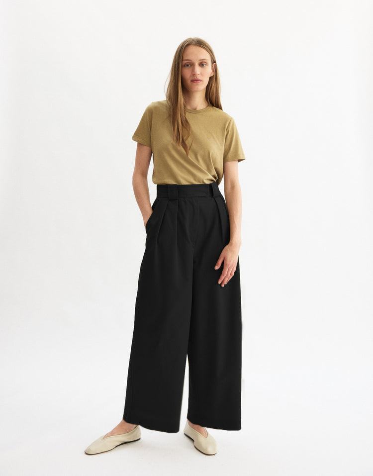A woman with long straight hair and a neutral expression stands against a white background wearing the Arlo Mott Grand Wide Leg Pant in Black, a tan short-sleeve t-shirt, and cream flat shoes.