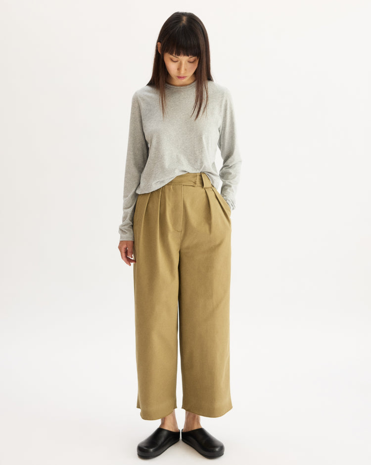 A woman stands against a white background, wearing a light gray long-sleeve top, Arlo Mott’s Grand Wide Leg Pant in Khaki, and black shoes. She looks down with her hands in her pockets.