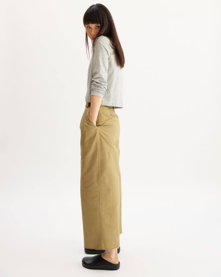 A woman with long dark hair stands sideways, looking over her shoulder. She wears a light gray long-sleeve top, Arlo Mott Grand Wide Leg Pant in Khaki with pockets, and black slip-on shoes against a white background.