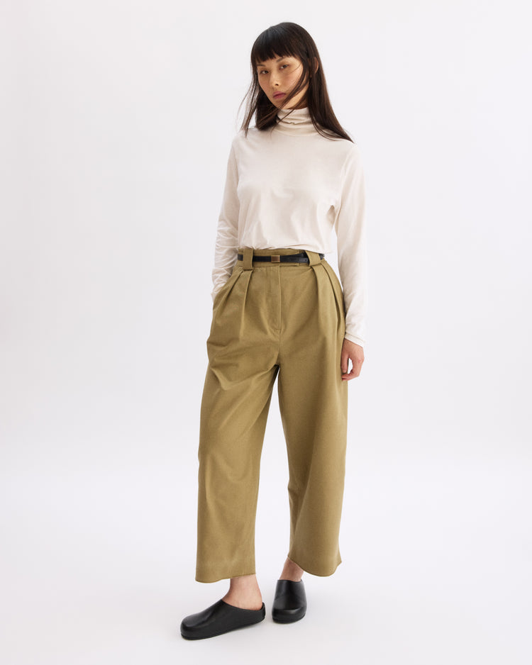 A person stands against a white background wearing a white turtleneck, Arlo Mott Grand Wide Leg Pant in Khaki with a black belt, and black slip-on shoes. Their hands are in their pockets and they have straight, long hair.