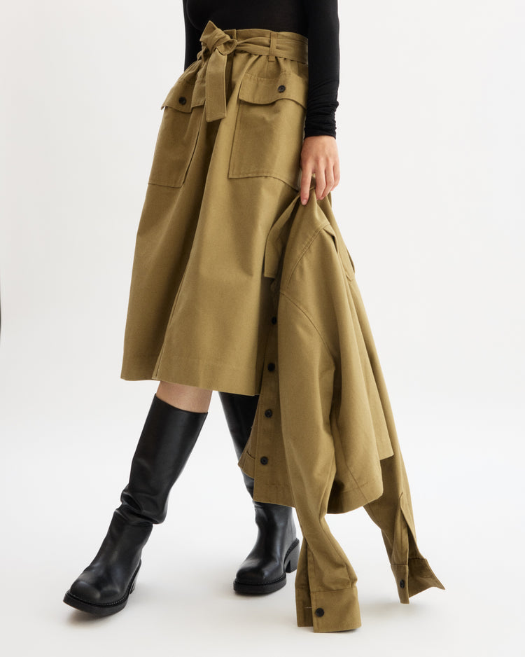 A person wears the Arlo Mott Felix Utility Midi Skirt in Khaki with a tie belt, black knee-high boots, and a black top, holding a matching khaki jacket against a plain white background.