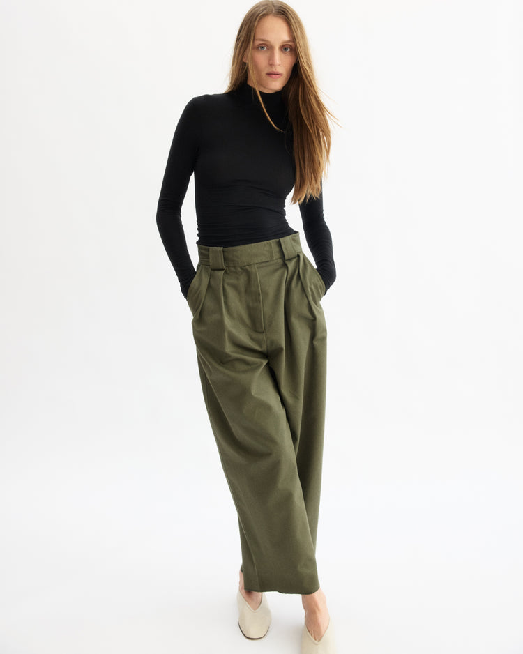 A woman stands against a white background wearing a black turtleneck, Arlo Mott’s Grand Wide Leg Pant in Olive, and beige shoes; her hands are in her pockets and her hair is down.