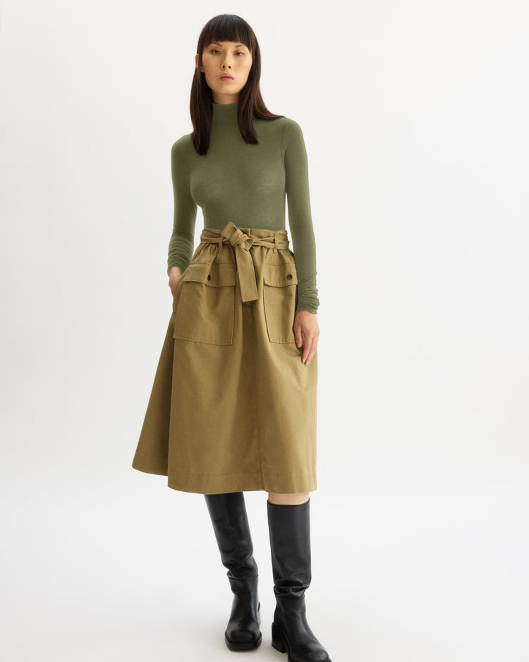 A woman poses against a white background in the Arlo Mott Chambers Long Sleeve Body Suit in Olive, paired with a high-waisted beige skirt featuring large front pockets and a tied belt, styled with knee-high black boots.