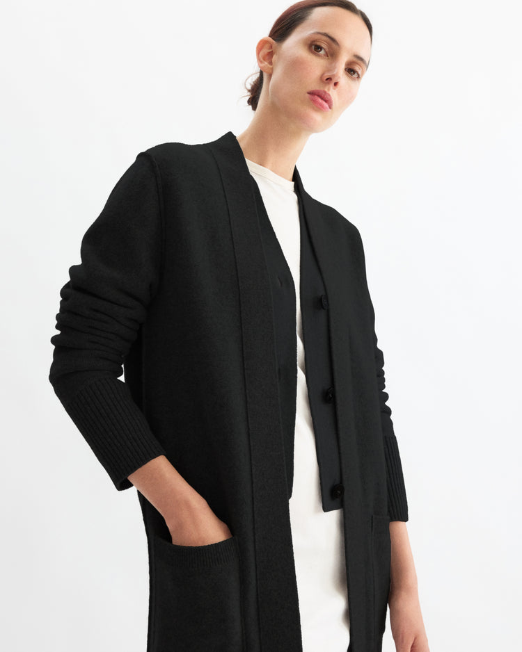 A person with pulled-back hair models the Arlo Mott Sidney Duster Cardigan in Black over a white top, standing against a plain white background with one hand in their pocket.