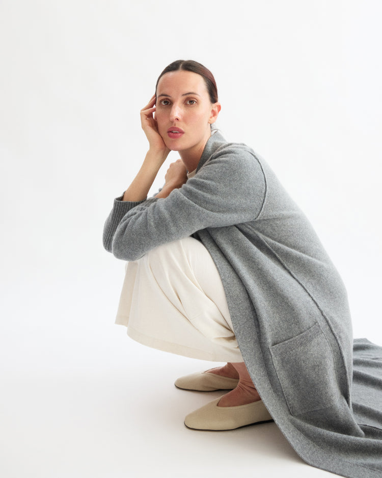 A woman with dark hair pulled back sits on her heels, wearing the Arlo Mott Sidney Duster Cardigan in Heather Grey, cream pants, and beige shoes, posing calmly against a plain white background.