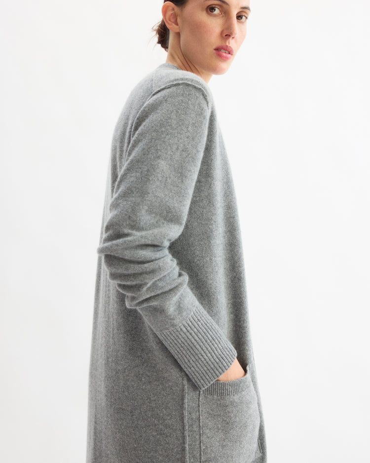 A person stands sideways against a white background, wearing the Arlo Mott Sidney Duster Cardigan in Heather Grey—a long wool and cashmere blend sweater with pockets—while looking over their shoulder.