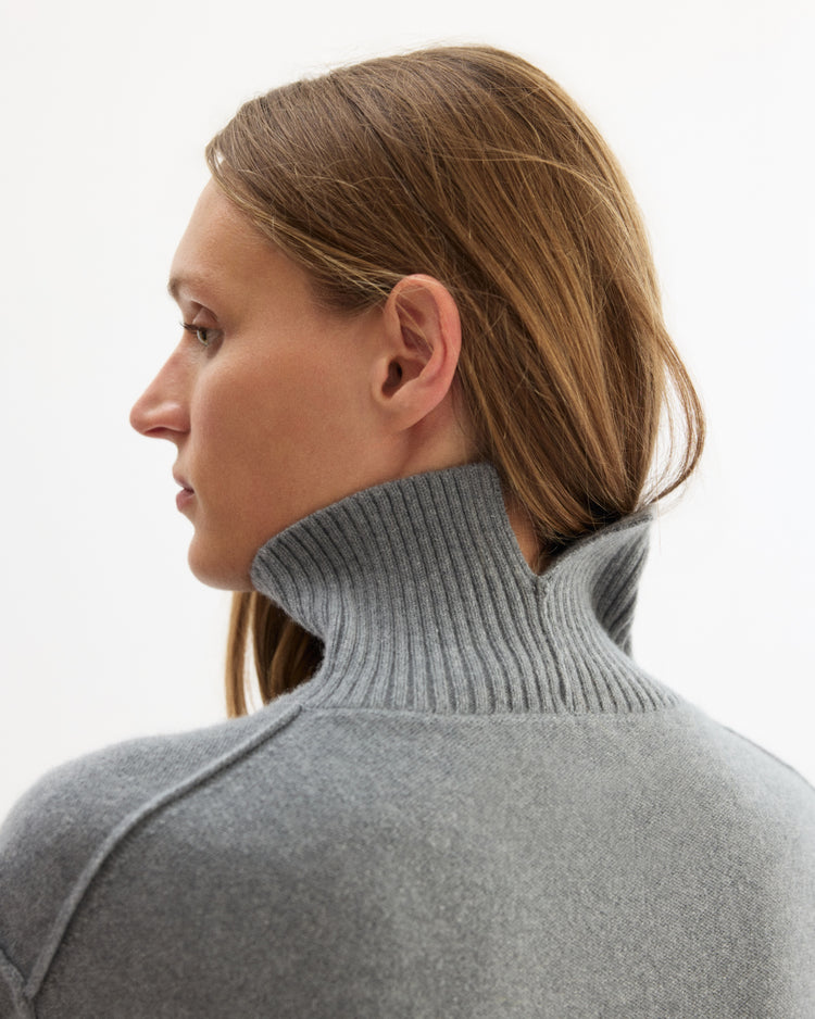 A woman with straight, light brown hair is shown in profile wearing the Arlo Mott Monroe Boxy Turtleneck in Heather Grey. She faces left against a plain white background.