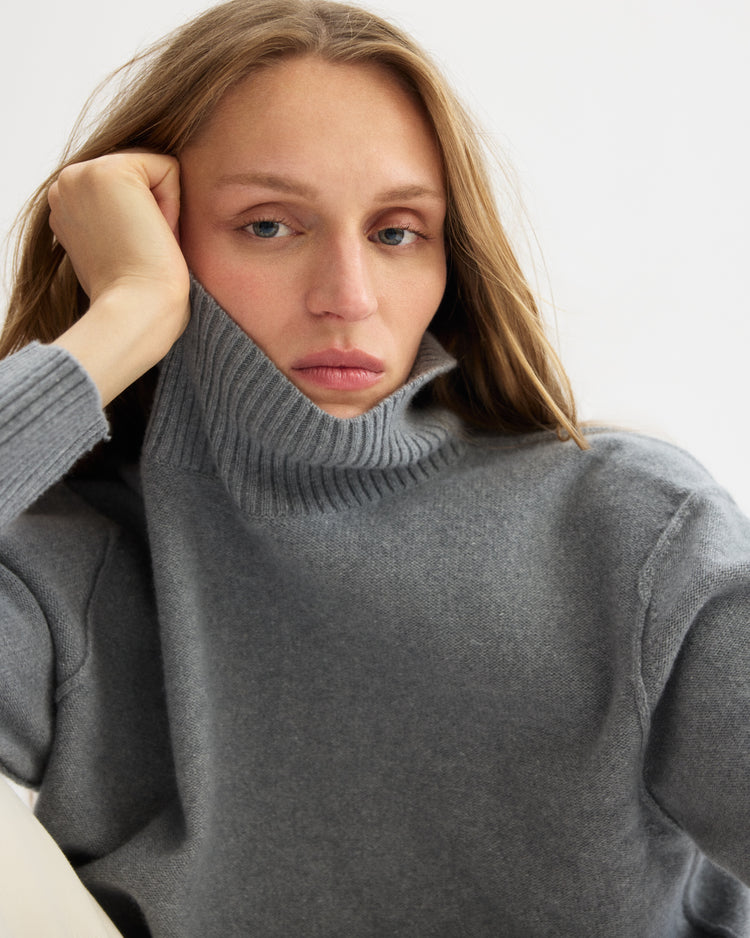 A person with long light brown hair wears the Arlo Mott Monroe Boxy Turtleneck in Heather Grey, resting their head on their hand and gazing thoughtfully at the camera against a plain background.