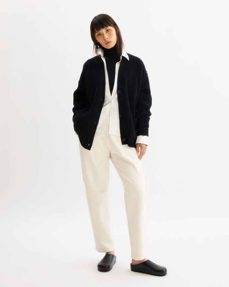 A woman with straight dark hair and bangs stands against a plain white background wearing Arlo Mott Baltic Barrel Leg Sweatpants in Chalk, a black cardigan over a white shirt, and black slip-on shoes, with her hands in her pockets.