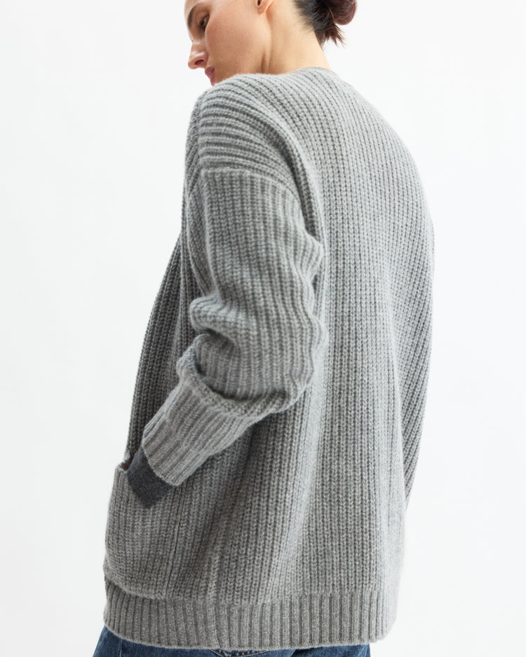 A person in the Arlo Mott Bedford Grandpa Cardigan in Heather Grey, paired with blue jeans, stands with their back to the camera and one hand in their pocket; hair is tied up against a plain white background.