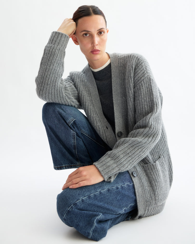 A person with dark hair pulled back wears the Arlo Mott Bedford Grandpa Cardigan in Heather Grey over a dark shirt and blue jeans, sitting with one knee raised and their elbow resting on it against a plain white background.