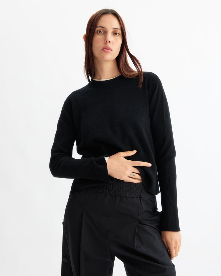 A person with long, straight brown hair wears the Arlo Mott Warren Classic Crew Neck in Black and black pants, standing against a plain white background with one hand resting across their midsection.