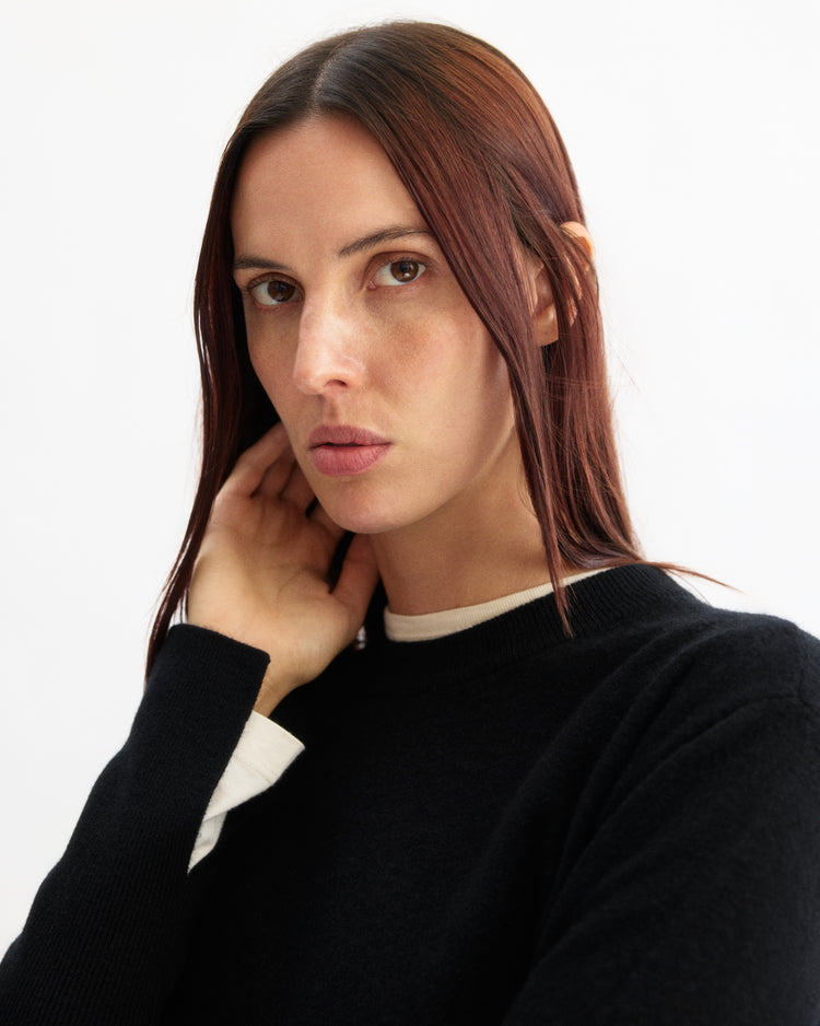 A person with long, straight brown hair and a neutral expression poses against a plain white background, wearing the Arlo Mott Warren Classic Crew Neck in Black over a white shirt, one hand touching the side of their neck.