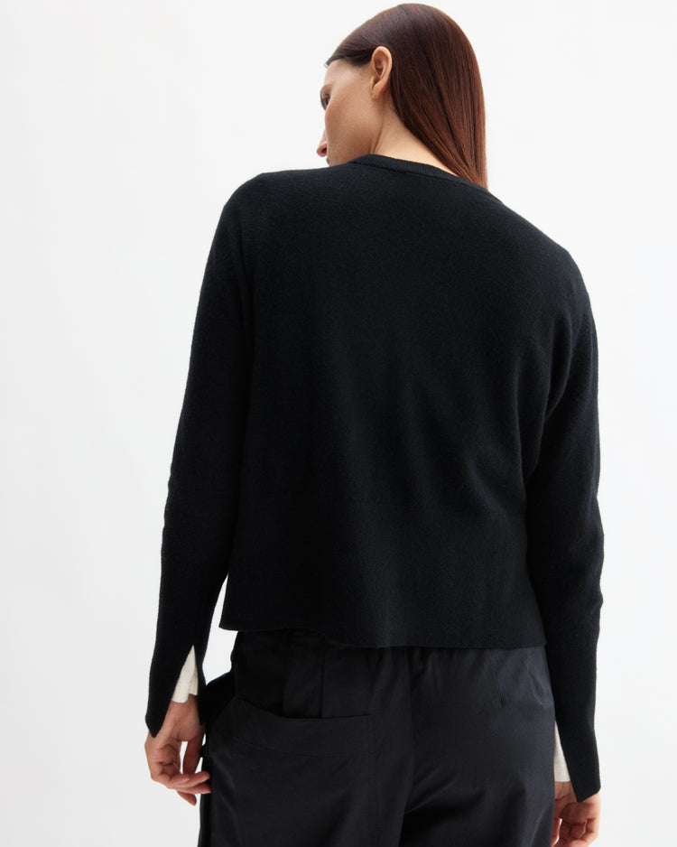 A person with long brown hair stands facing away, wearing the Arlo Mott Warren Classic Crew Neck in Black and black pants against a plain white background.