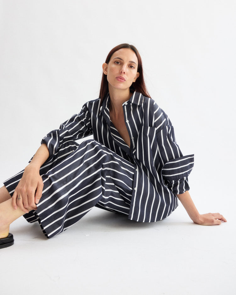 A woman with long brown hair sits on the floor against a white background, wearing Arlo Mott's Hudson Pull-on Pants in Navy Stripe, a wide-leg style made from soft Portuguese cotton, with one hand on the floor and the other on her knee.