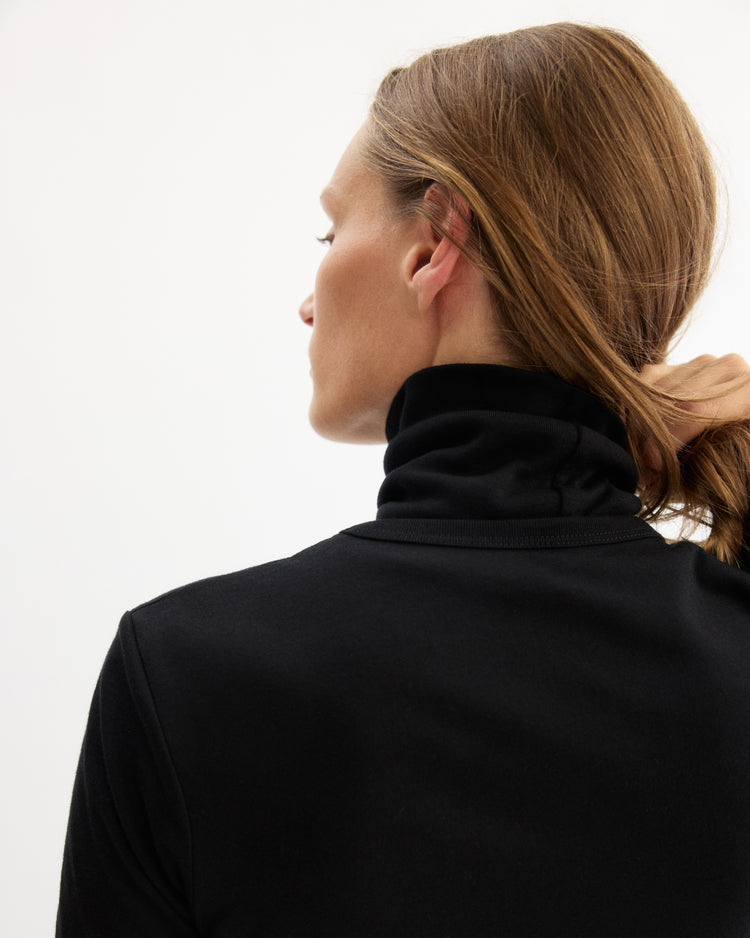 A person with light brown hair, seen from behind and in profile against a plain white background, gently holds their hair while wearing the Arlo Mott Prince Long Sleeve Turtleneck in Black.
