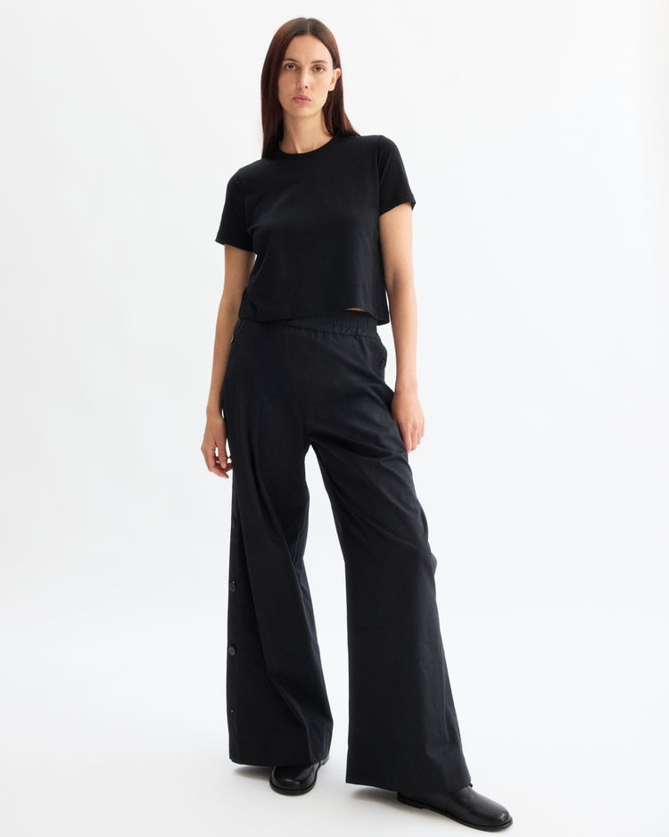 A woman with straight brown hair and a neutral expression stands against a white background, wearing the Arlo Mott Willow Short Sleeve Tee in Black, loose black pants with side buttons, and black shoes.
