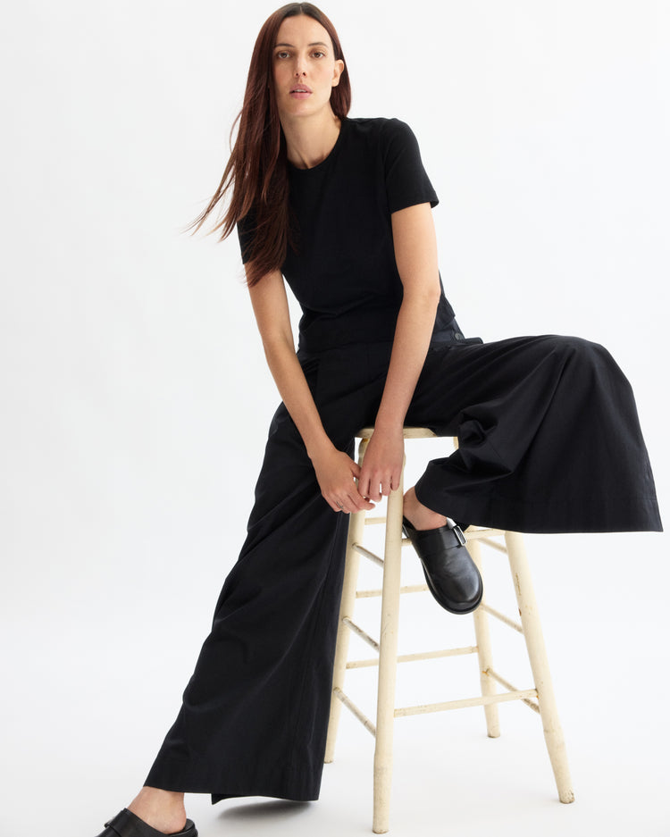 A woman with long, straight brown hair sits on a wooden stool wearing the Arlo Mott Willow Short Sleeve Tee in Black with wide-leg black pants and black shoes, set against a plain white background.