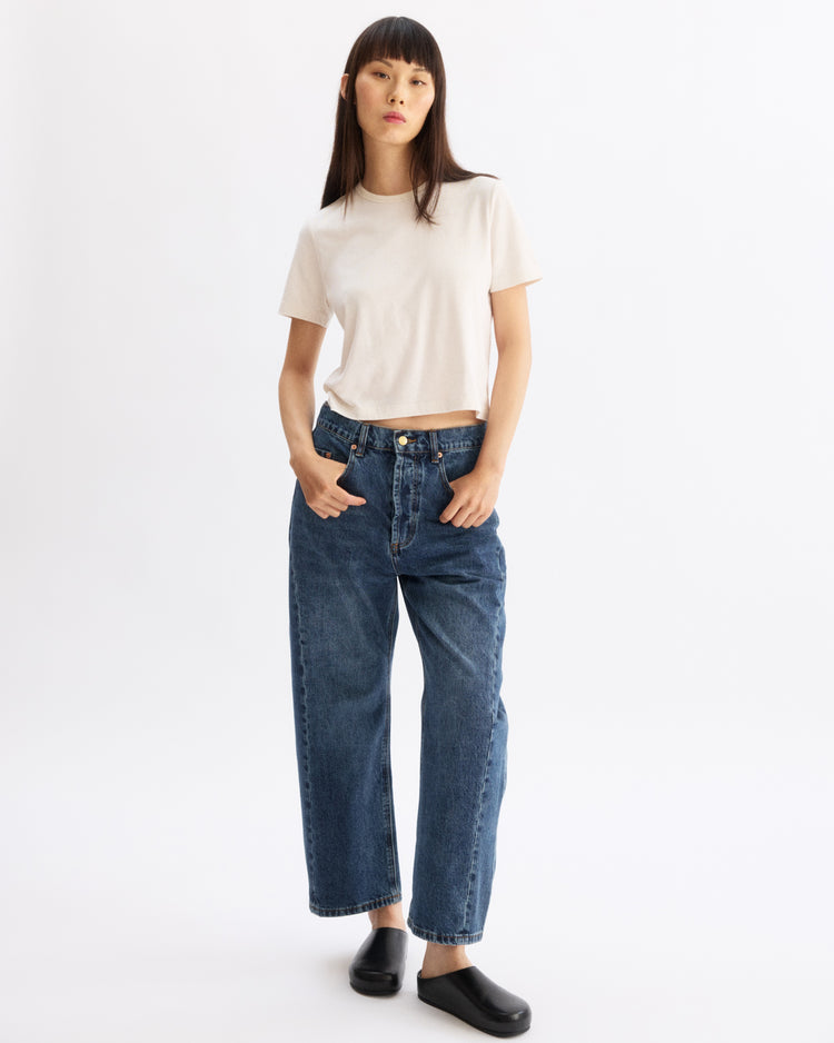 A woman stands against a white background wearing the Arlo Mott Willow Short Sleeve Tee in Chalk, high-waisted relaxed blue jeans, and black slip-on shoes, with her hands in her pockets and a neutral expression.
