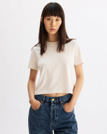 A person with straight dark hair and bangs stands against a plain background, wearing the Arlo Mott Willow Short Sleeve Tee in Chalk and blue jeans, hands in pockets, looking confidently at the camera.