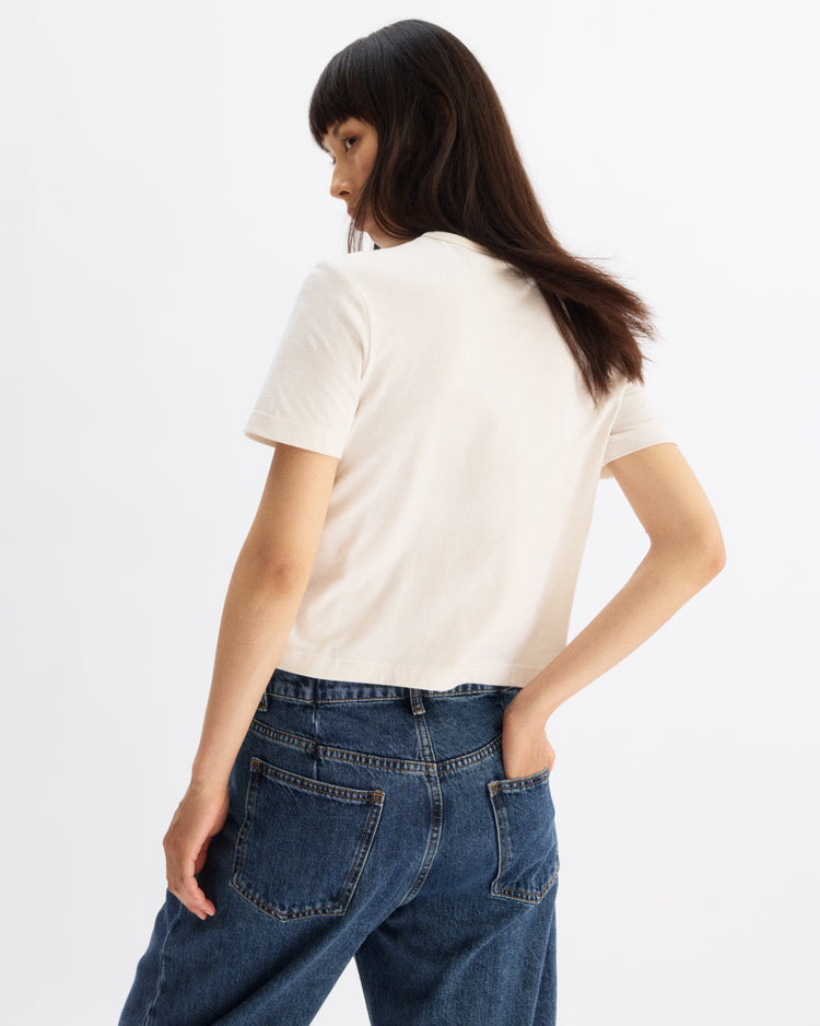 A woman with long dark hair stands sideways, looking over her shoulder. She wears the Arlo Mott Willow Short Sleeve Tee in Chalk and blue jeans, one hand in her back pocket, against a plain white background.