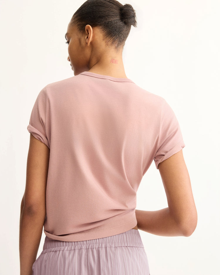 A woman with a neat bun and a small red neck tattoo faces away, wearing the Arlo Mott Willow Short Sleeve Tee in Dusty Rose and mauve pleated pants.