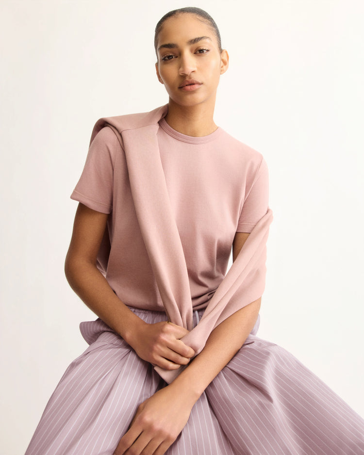 A person with slicked-back hair wears the Arlo Mott Willow Short Sleeve Tee in Dusty Rose, with a matching sweater draped over their shoulders. They sit confidently and look at the camera against a light, neutral background.