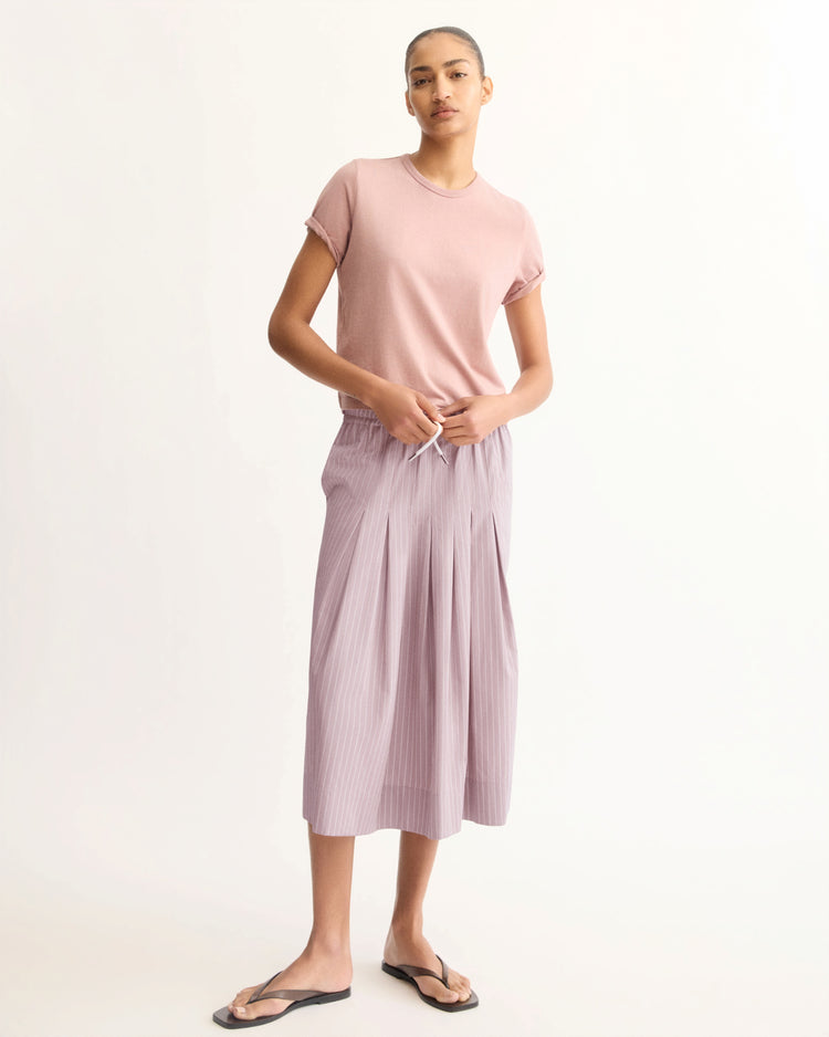 A person stands against a plain background wearing a light pink short-sleeve shirt, the Arlo Mott Gates Full Midi Skirt in Rose Stripe, black flip-flops, and has their hair pulled back.
