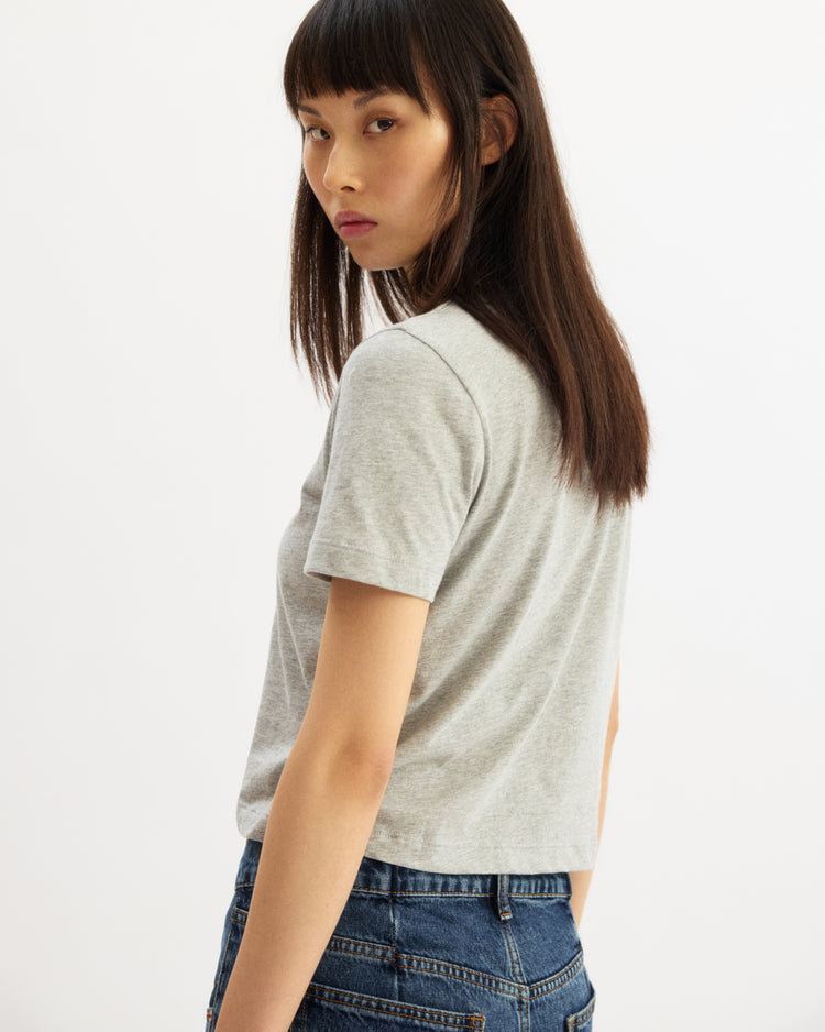 A young woman with straight, dark brown hair wears the Arlo Mott Willow Short Sleeve Tee in Heather Grey and blue jeans, standing sideways and looking over her shoulder with a neutral expression against a plain background.