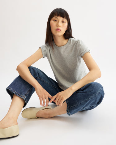 A woman with straight dark hair sits on the floor, wearing the Arlo Mott Willow Short Sleeve Tee in Heather Grey, blue jeans, and beige slip-on shoes, looking confidently at the camera against a plain white background.