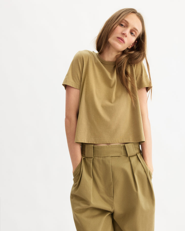 A woman wears the Arlo Mott Willow Short Sleeve Tee in Khaki, a boxy fit made from soft Supima cotton, paired with high-waisted pleated khaki trousers. She stands against a white background, hands in pockets, gazing slightly to the side.